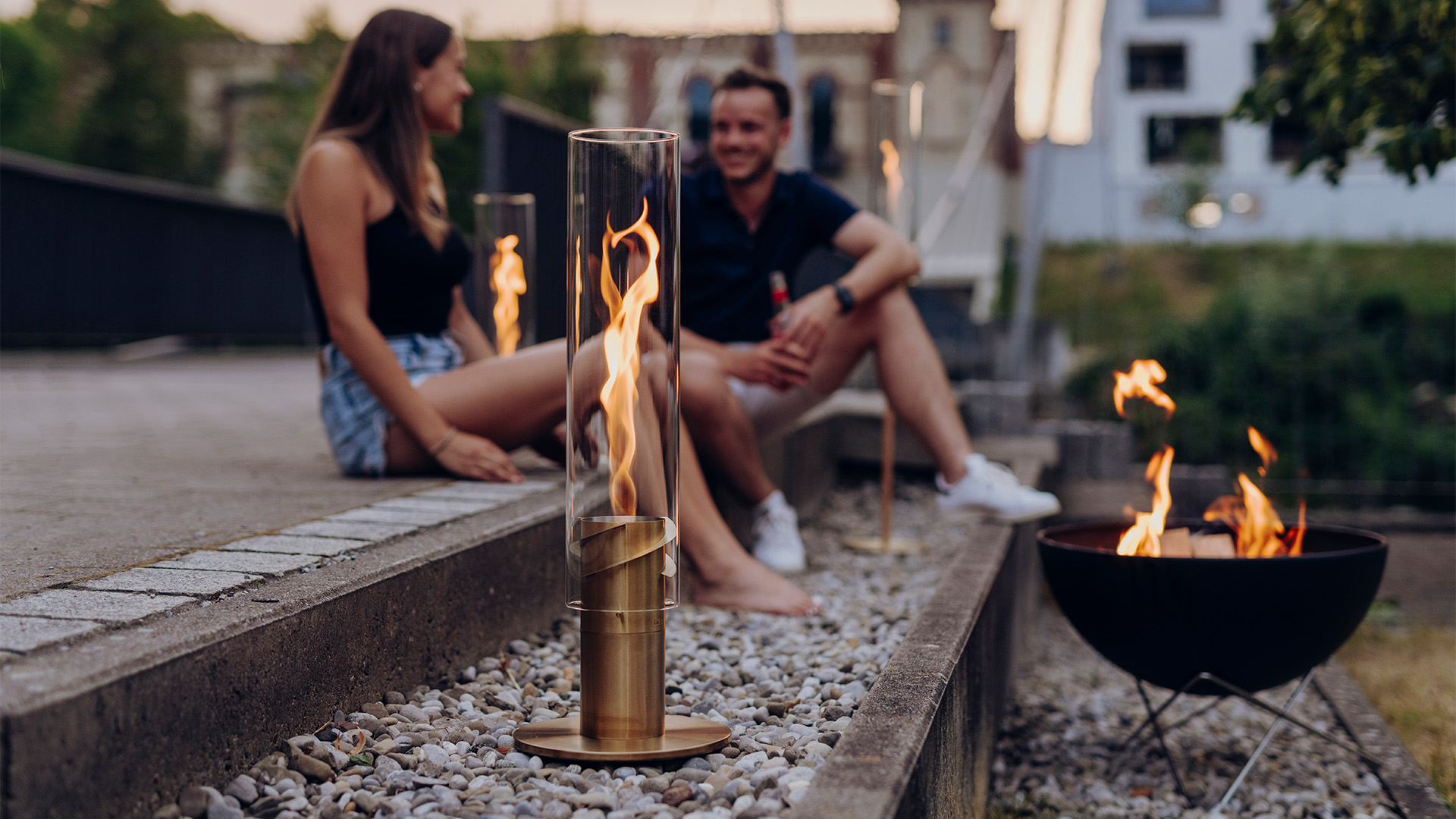 Scene showing a couple sitting on a bench next to a fire pit, where the Spin elevation Gold stands out for its practical elegance.