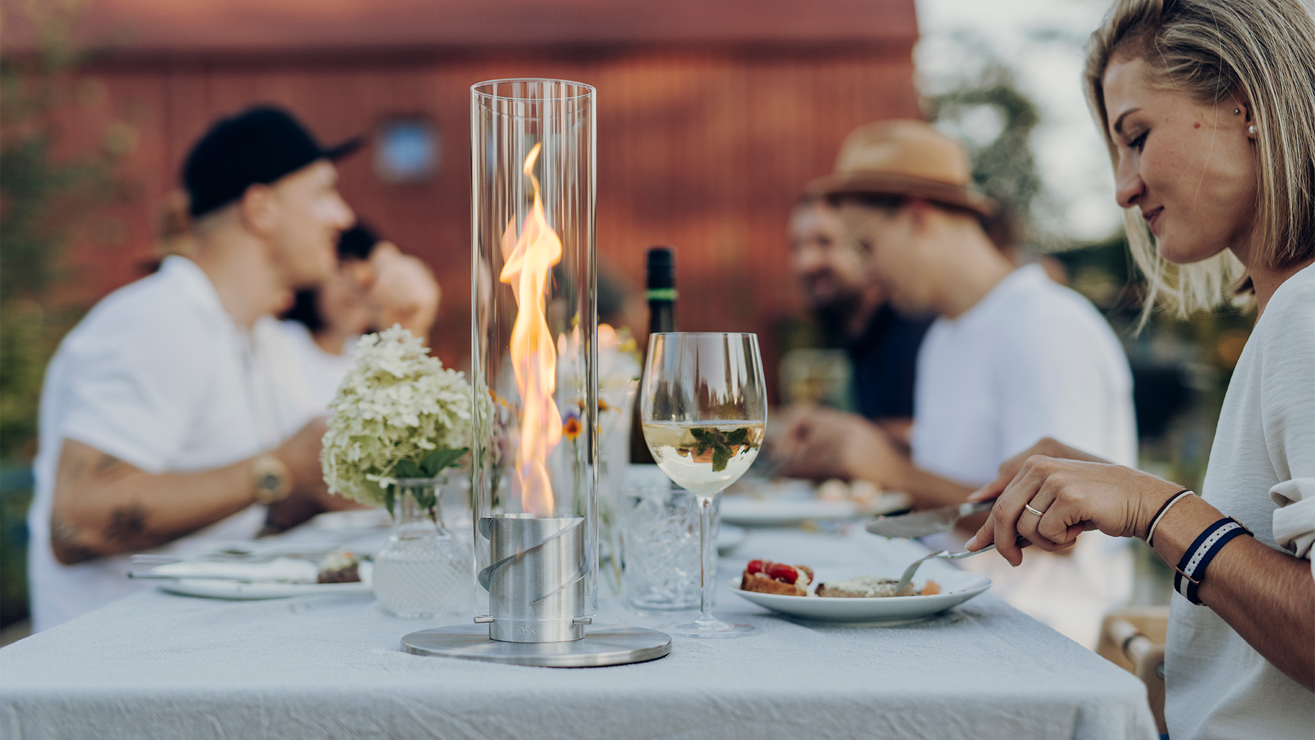 The photo presents a woman sitting at a table with a plate of food, with the SPIN 900 Tabletop Fireplace silver.