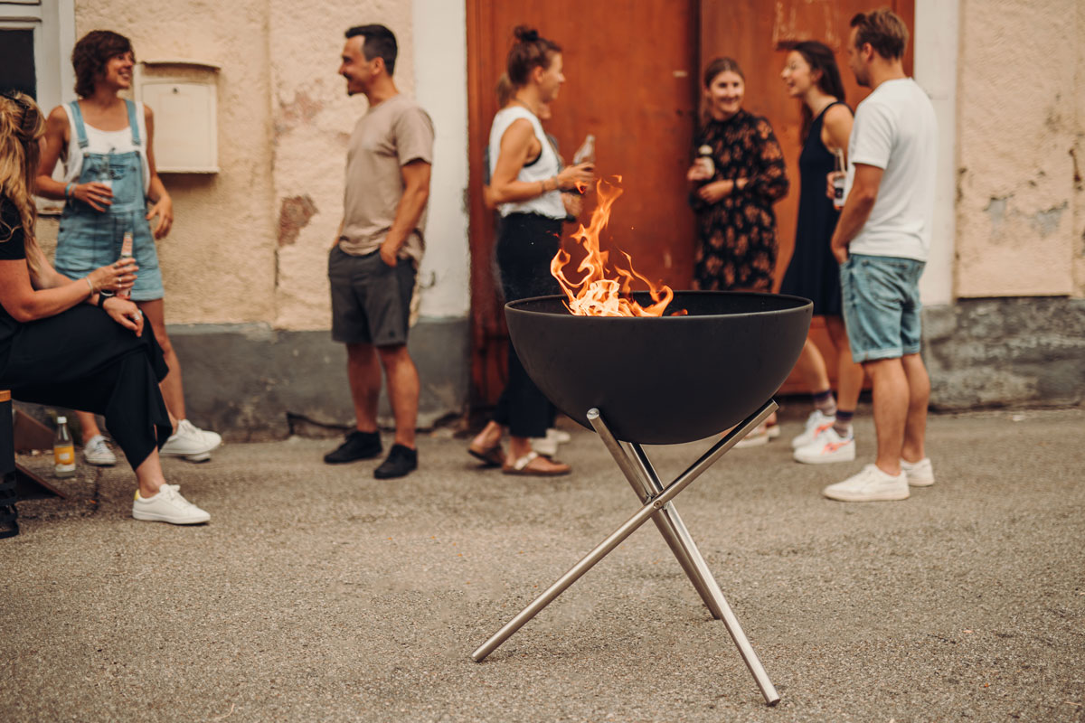 BOWL 70 Fire Bowl with tripod Scene showing a group of people standing around a fire pit, where the 00258 Bowl 70 with Tripod stands out for its practical elegance.