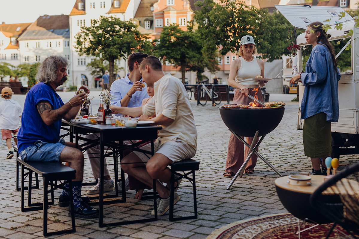 The Bowl 70 BBQ Set High is shown as a group of people sitting around a table outside, creating a stylish and accessible visual.