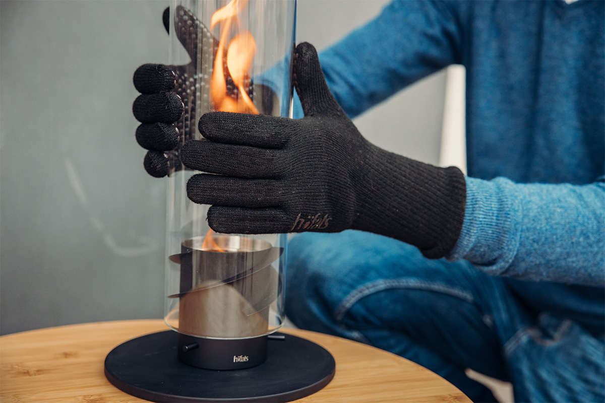 Fire Gloves Aramid This image shows a person in a blue sweater and black gloves holding a tabletop fireplace with a fire inside, with the Fire Gloves Aramid.
