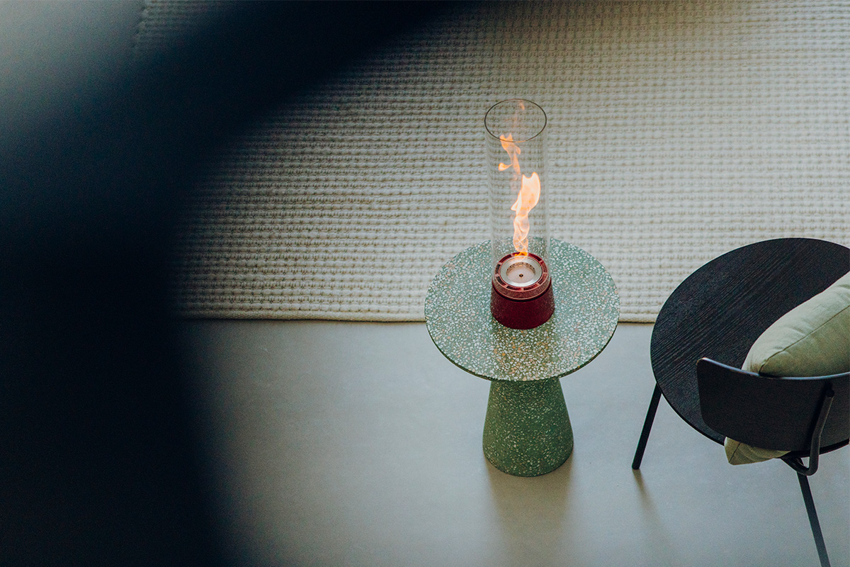 SPIN air 1200 Tabletop Fireplace enamel red A depiction of a chair and a table fire in a room, featuring the 00814 Spin Air 1200 Tablefire Emaille red as a central and purposeful element.