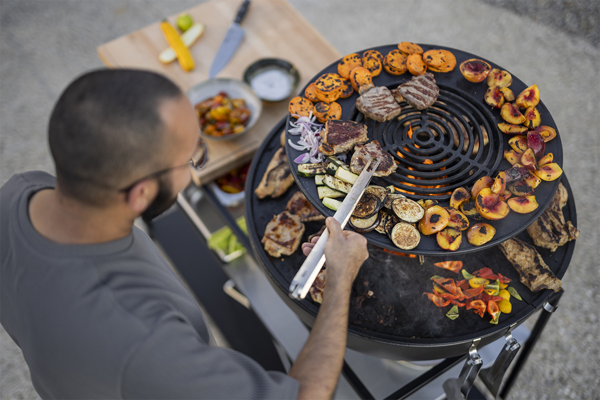 The 00617 Bowl 70 Warming Ring is shown as a man is grilling on a large grill with a tong, creating a stylish and accessible visual.