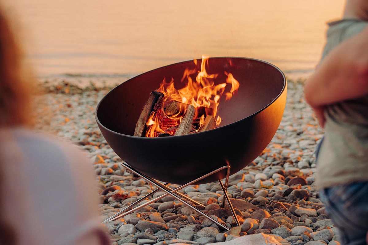 A woman sitting in front of a fire pit, with the 00249 Bowl 57 with starstand adding a functional and elegant touch to the scene.