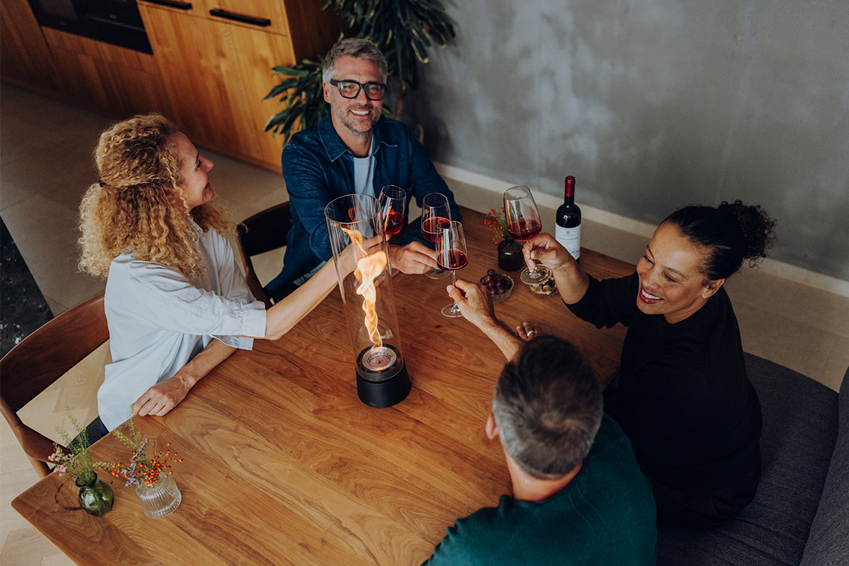 SPIN air 1200 Tabletop Fireplace matte black SPIN air 1200 und 900 mattschwarz auf einem Küchentisch mit vier Personen welche mit einem Rotwein anstoßen