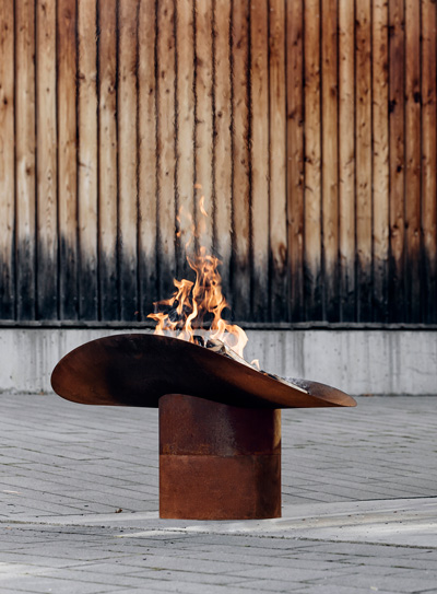 A view of a fire pit with a wooden fence behind it, with the ELLIPSE Fire Bowl.